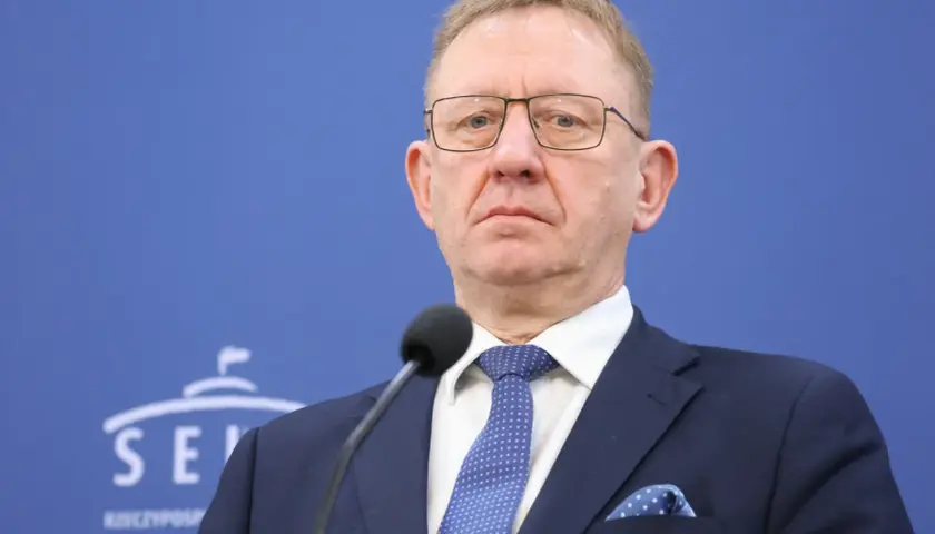 Mężczyzna w okularach i granatowym garniturze przed mikrofonem na tle niebieskiej ściany z logo Senatu.