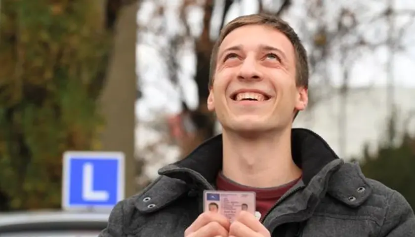 Szczęśliwy młody mężczyzna trzyma prawo jazdy, sprawdzając czy jest do odbioru. W tle niebieski znak "L".