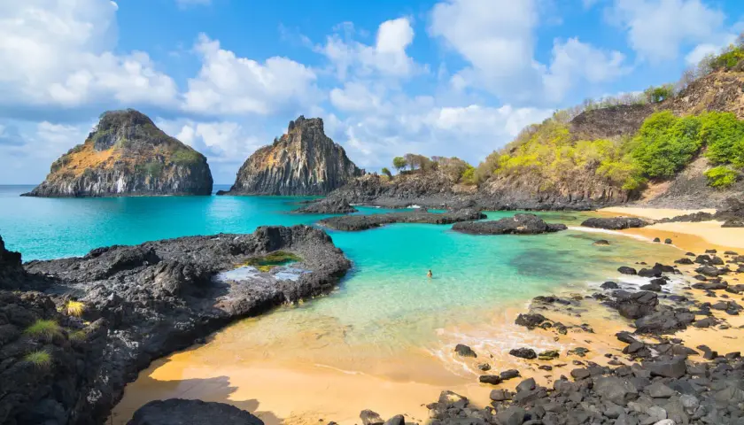 Rajska plaża na Fernando de Noronha z turkusową wodą, skalistymi formacjami i samotną postacią kąpiącą się w oceanie.