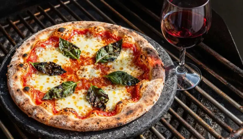 Pizza mit langer Gehzeit auf dem Grill, belegt mit Tomaten, Mozzarella und Basilikum, daneben ein Glas Rotwein.