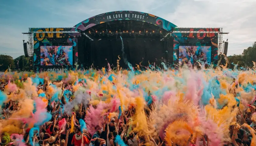 Une foule en liesse lance de la poudre colorée pour créer un festival de musique inoubliable. L'ambiance est électrique sous la scène "IN LOVE WE TRUST".