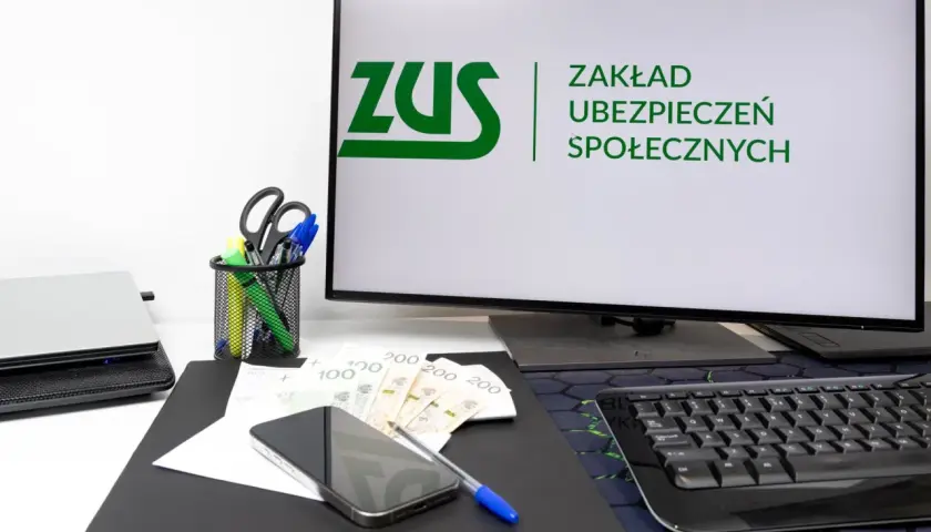 Biurko z monitorem wyświetlającym logo ZUS, pieniędzmi, telefonem i artykułami biurowymi.