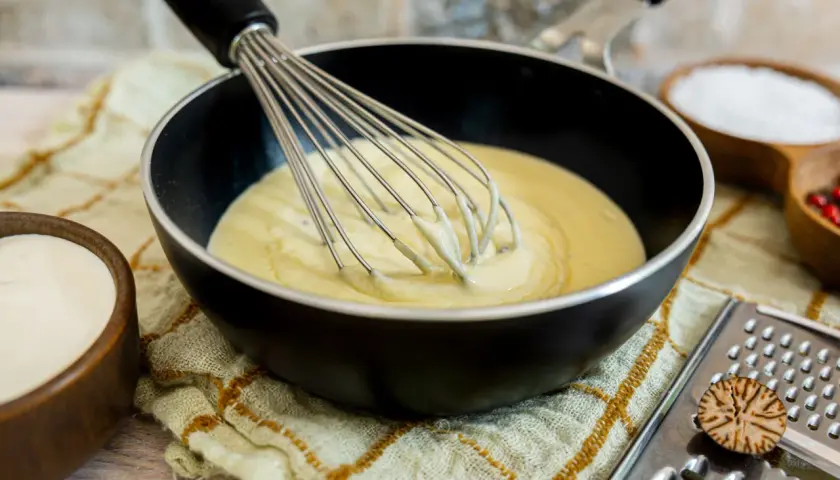 Eine cremige low carb bechamel wird in einer Pfanne mit einem Schneebesen verrührt. Daneben stehen Schüsseln mit Sahne, Salz und Pfefferkörnern.