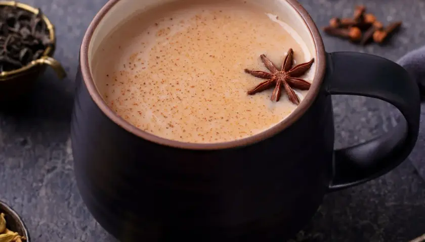 Aromaticzny napój, idealny na chłodne dni. Gorący **masala chai** w czarnej filiżance, ozdobiony anyżem. Obok liście herbaty, kardamon i goździki.