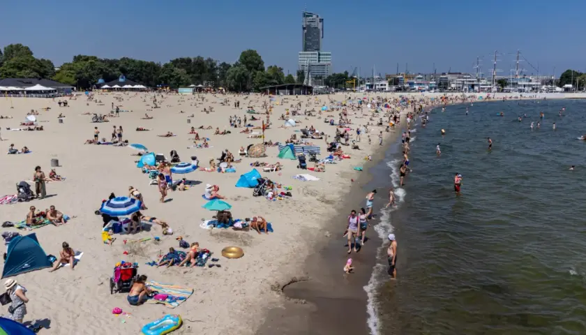 Słoneczna plaża w jednej z miejscowości koło Gdyni, pełna wypoczywających ludzi. W tle widać drapacz chmur i żaglowce.