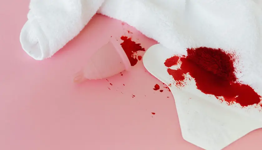 Sennik krew menstruacyjna: kubeczek i podpaska z plamami krwi na różowym tle.