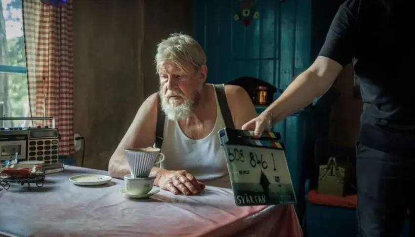Rolf Lassgård sitter vid ett bord med en kaffebryggare. En person håller upp en filmklappa för en ny serie med Rolf Lassgård.