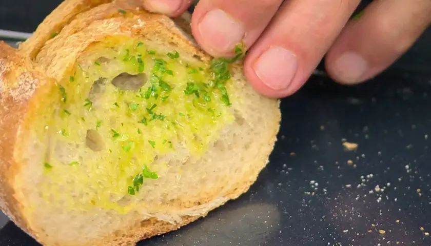 Un trozo de pan rústico con una generosa capa de mantequilla de ajo y perejil picado, listo para ser horneado.