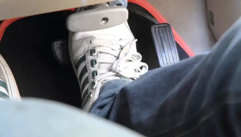 Un pied en basket Adidas appuie sur la pédale de frein, qui semble un peu molle. Le tapis de sol est noir avec un contour rouge.