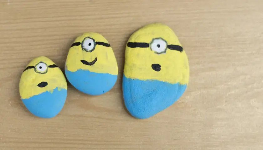 Trzy kamienie polne pomalowane na żółto i niebiesko, przypominające Minionki. Zabawne malowanie kamieni polnych.