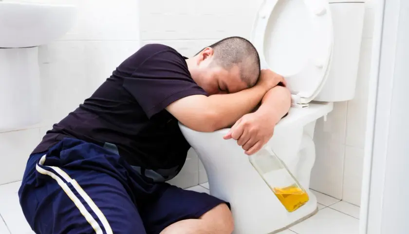 Mężczyzna śpi na muszli klozetowej, trzymając butelkę. Widać, że doświadczył wymiotów po alkoholu.
