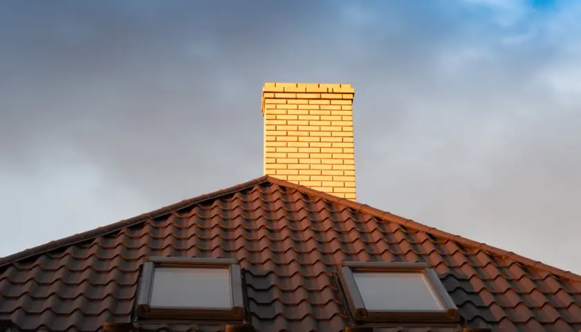 Jasna rura kominowa ceramiczna na ciemnym dachu z dachówkami, pod niebo z chmurami.