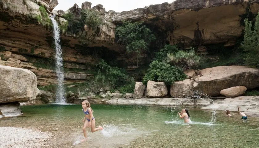 Niños disfrutan en una **piscina natural** bajo una cascada, rodeada de rocas y vegetación.
