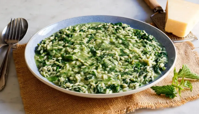 Un cremoso risotto con ortiche, servito in un piatto blu, con forchetta e coltello a lato. Un pezzo di parmigiano e una grattugia completano la scena.