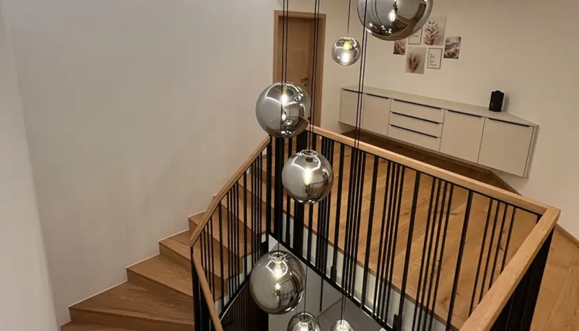 Nowoczesna klatka schodowa z drewnianymi stopniami i czarną balustradą, ozdobiona wiszącymi kulistymi lampami.