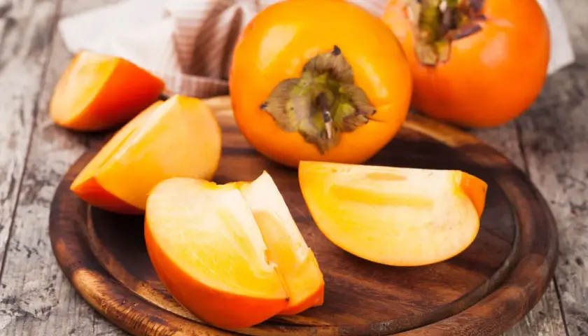 Pomarańczowe owoce kaki, całe i pokrojone w ćwiartki, na drewnianej desce. Odkryj owoc kaki właściwości zdrowotne.