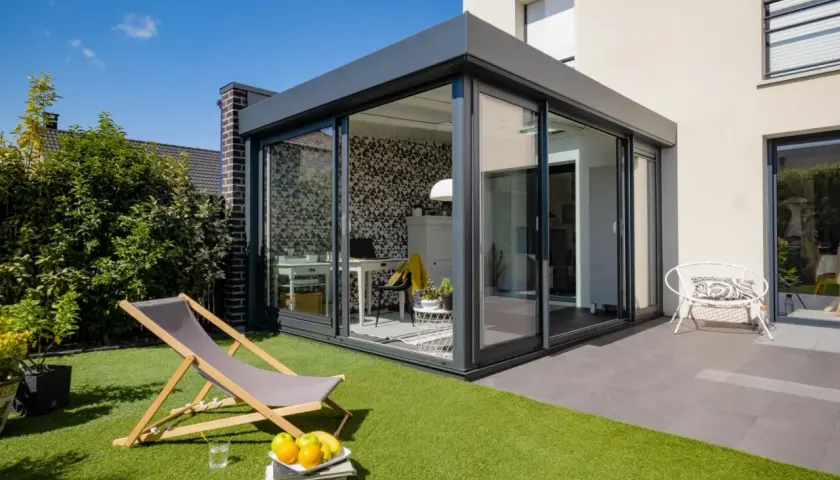 Aménagement véranda 15m2 moderne avec bureau et espace détente extérieur. Chaise longue, fruits et fauteuil blanc invitent au repos.