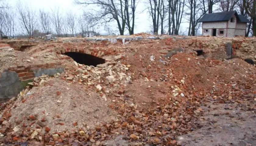 Ruiny ceglane z łukiem i mały domek w tle, otoczone drzewami.