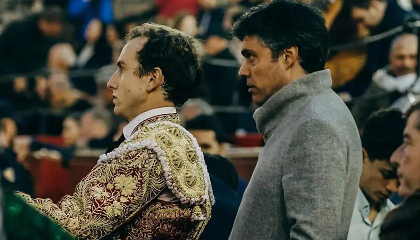 Un torero con traje de luces bordado y zapatillas torero, junto a un hombre con abrigo gris, observan la plaza.