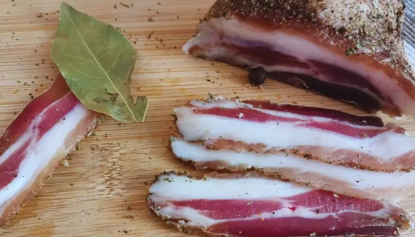 Pancetta co to? Pokrojony w plastry, przyprawiony boczek na desce. Obok liść laurowy i kawałek większego kawałka.
