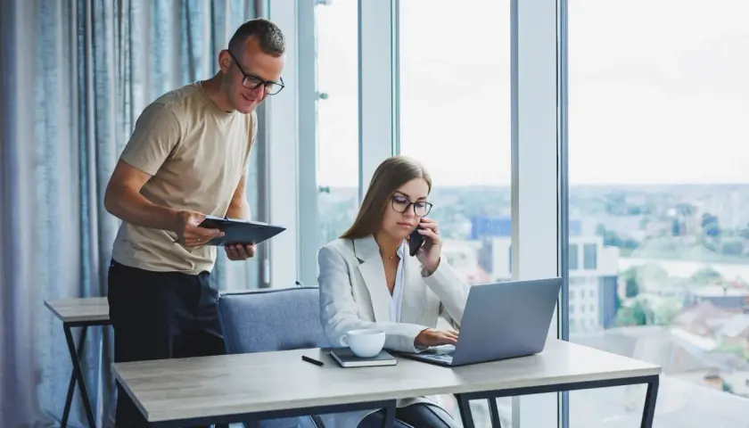 Un homme consulte un document pendant qu'une femme au téléphone travaille sur son ordinateur portable, illustrant l'efficacité de l'e-commerce B2B.