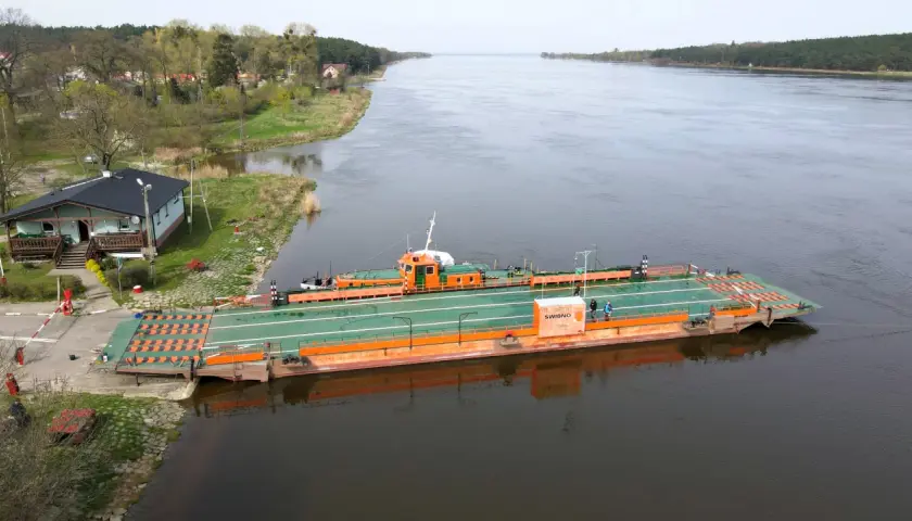 Prom "Świbno" czeka na pasażerów przy brzegu, gotowy do przeprawy przez szeroką rzekę.