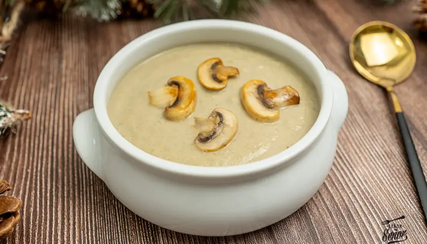 Crema de champiñones servida en un cuenco blanco, decorada con rodajas de champiñones salteados. Una cuchara dorada descansa a la derecha.
