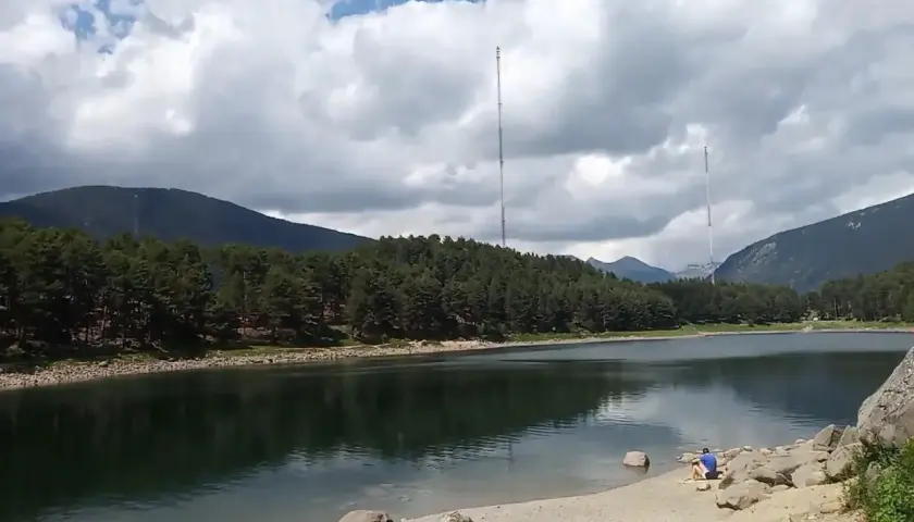 Persona sentada en la orilla del lago de Engolasters, rodeado de pinos y montañas, con dos torres de telecomunicaciones al fondo.