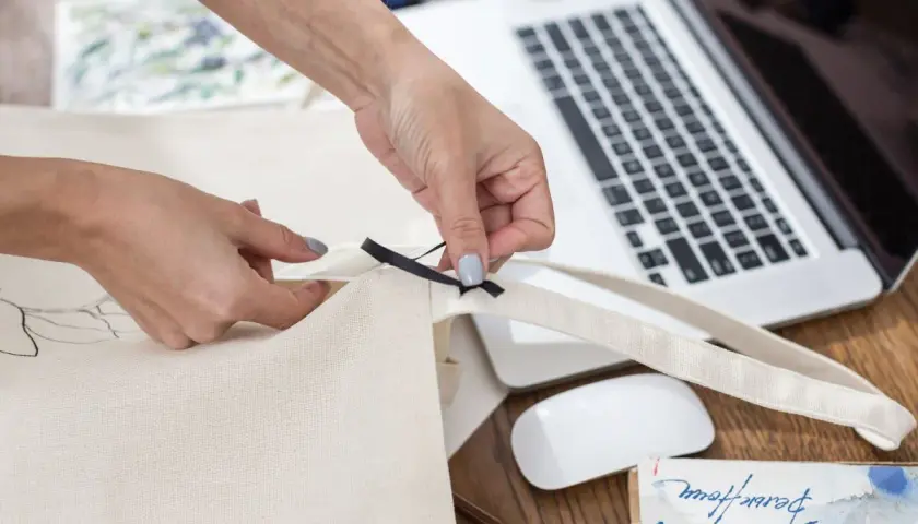 Ręce szyją torbę shopper, dodając czarną wstążkę. Na biurku laptop, myszka i materiały do szycia.