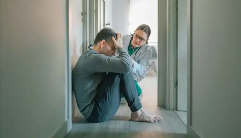 Lekarz wspiera pacjenta zmagającego się ze splątaniem w psychiatrii. Mężczyzna siedzi na podłodze, przygnębiony, a lekarka pochyla się nad nim z troską.