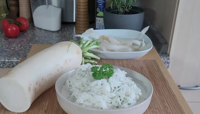 Ein frischer Rettich liegt neben einer Schüssel mit cremigem Rettichsalat, garniert mit Petersilie. Perfekt für ein leichtes Rettichsalat Rezept.