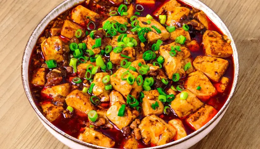 En skål med kryddig mapo tofu, toppad med färsk salladslök, serverad på ett träbord.