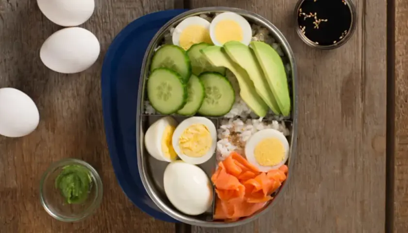 A bento box filled with rice, sliced cucumber, avocado, smoked salmon, and hard-boiled egg.