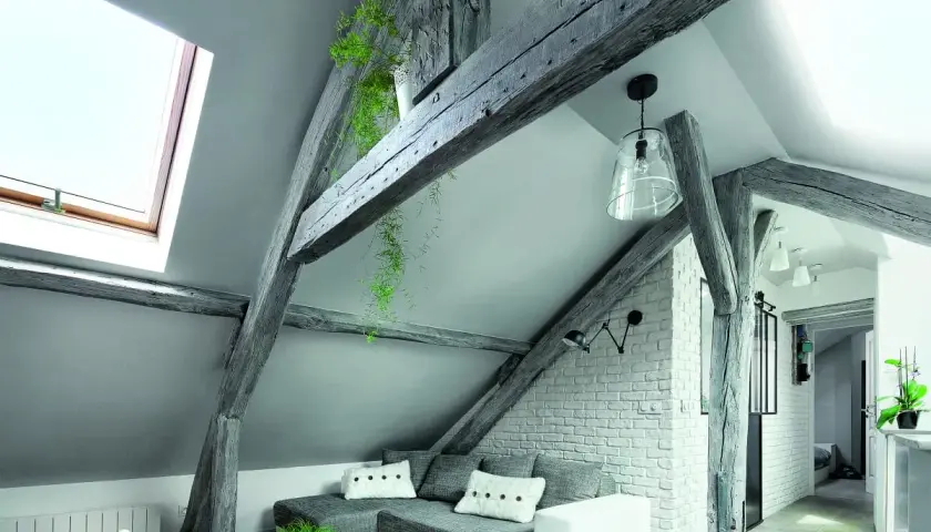 Przytulna mansarda z widocznymi belkami stropowymi, szarą kanapą i oknem dachowym.