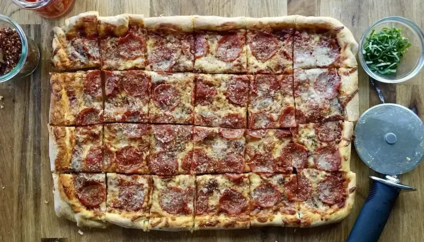 Prosty przepis na pizzę: prostokątna pizza pepperoni z serem, pokrojona na kwadraty, obok słoiczki z dodatkami i krajalnica.
