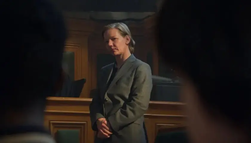 Sandra Hüller, con traje gris, se mantiene firme en una sala de audiencias, con las manos entrelazadas, mientras la cámara se enfoca en su rostro pensativo.