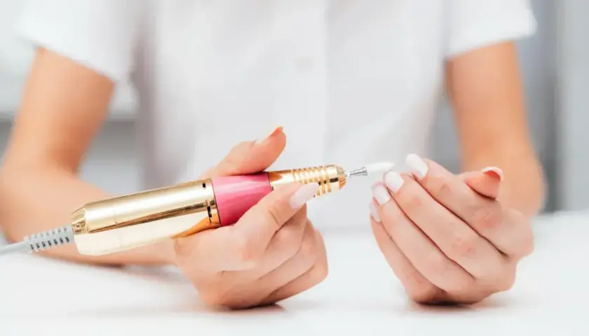 Dłonie z białymi paznokciami trzymają różowo-złotą frezarkę. Dowiedz się, jak używać frezarki do paznokci, aby uzyskać perfekcyjny manicure.