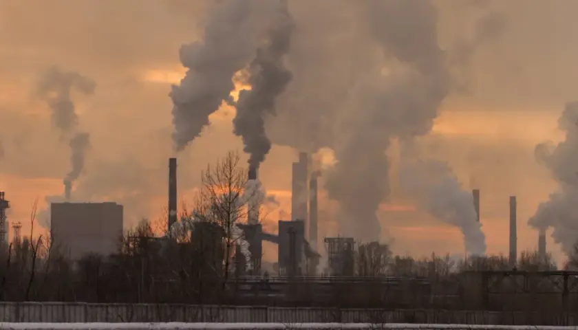 Chimeneas industriales expulsan humo denso, creando un paisaje sombrío. Las partículas contaminantes se elevan, tiñendo el cielo de un naranja opaco.