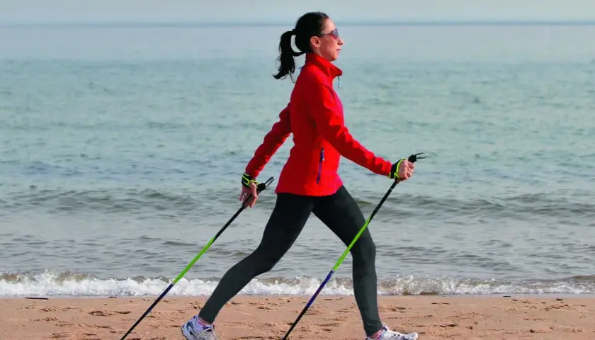 Kobieta w czerwonej kurtce ćwiczy marsz z kijkami na plaży. Pokazuje, jak chodzić z kijkami, wykorzystując je do aktywności fizycznej nad morzem.