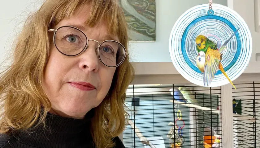 Aase Berg med glasögon tittar mot kameran, med en fågelbur och en teckning av två undulater i bakgrunden.