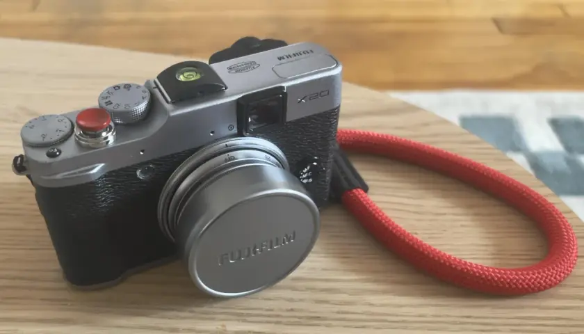 Srebrny aparat Fujifilm X20 z czerwoną smyczą leży na drewnianym blacie.