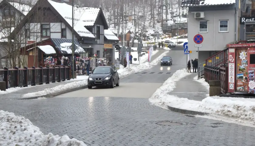 Zimowy deptak w Szklarskiej Porębie z samochodami na ulicy i sklepikami z pamiątkami.