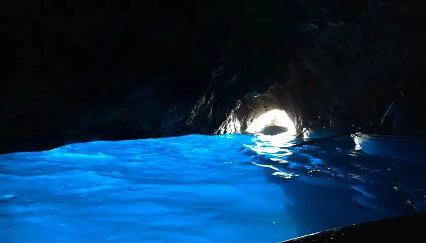 Acqua azzurra e luminosa in una grotta buia, con un'uscita illuminata dove si intravede una barca.