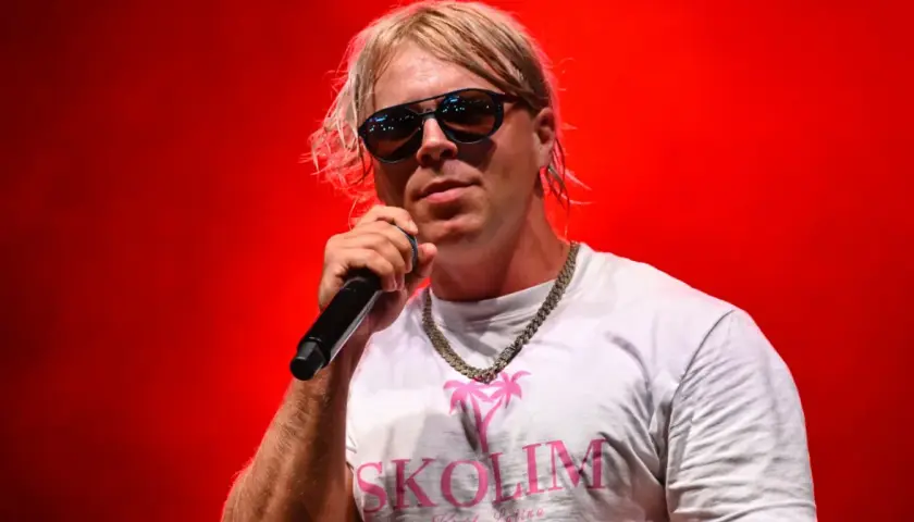 Skolim w okularach i białej koszulce z napisem "SKOLIM" śpiewa na czerwonym tle. Zastanawiasz się, ile Skolim bierze za koncert?