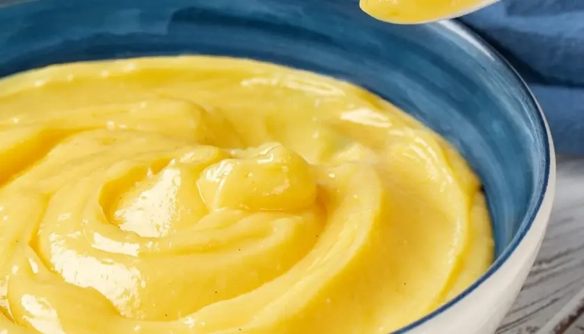 Una ciotola blu piena di deliziosa crema pasticcera vegana, con un cucchiaio che ne preleva una porzione.