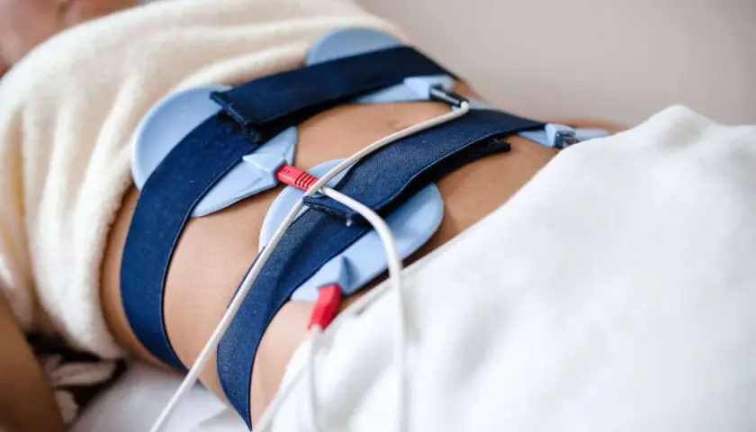 Osoba leży pod kocem z pasami i elektrodami na brzuchu, przygotowana do zabiegu ems trening.