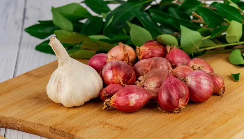 Knoblauch und Schalotten auf einem Holzbrett, bereit als Hausmittel gegen Ohrenschmerzen. Grüne Blätter im Hintergrund.