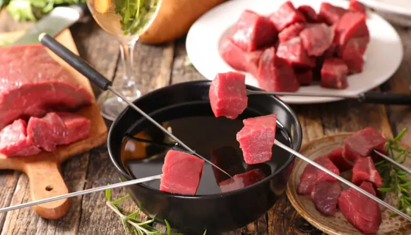 Préparer une fondue bourguignonne : des dés de bœuf crus sur des brochettes, prêts à être plongés dans l'huile chaude. Un verre de vin blanc à côté.