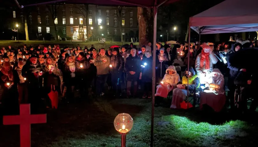 Menschen versammeln sich bei Kerzenlicht zu einem Gottesdienst am Heiligabend vor einem Gebäude. Ein rotes Kreuz steht im Vordergrund.