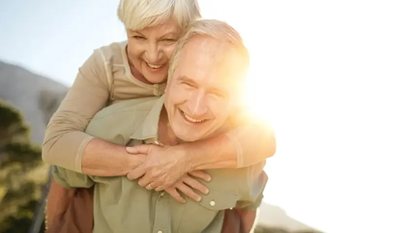 Ein Mann trägt eine Frau auf dem Rücken. Beide lachen und genießen die Sonne. Ein Bild von wahrer **Liebe im Alter**.